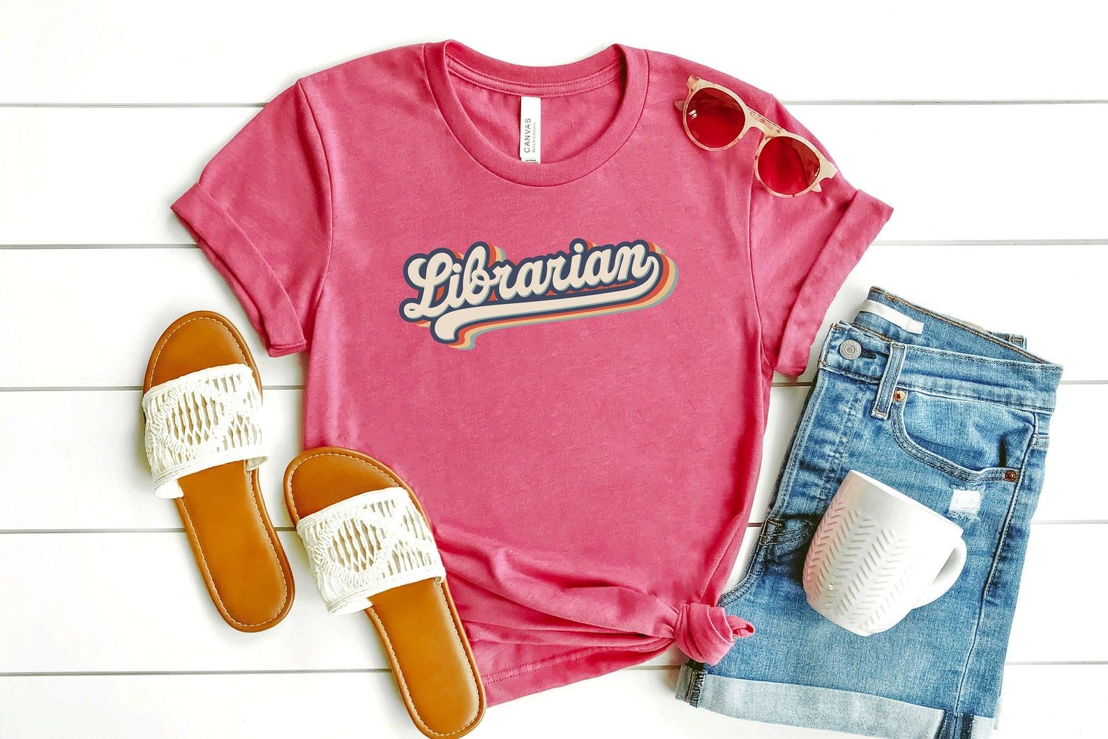 Image of a pink t-shirt reading "librarian" in colorful font. Beside the shirt are shorts, sandals, a coffee mug, and sunglasses. 