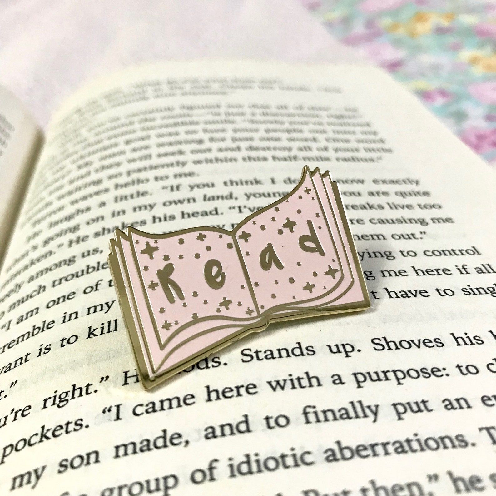 Pink enamel pin sitting on an open book. It reads "read," in gold letters. 