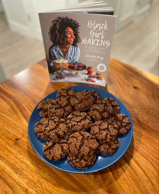 Crinkled cocoa gingersnap cookies covered in coarse sugar on a blue plate, alongside Jerrelle Guy's cookbook Black Girl Baking
