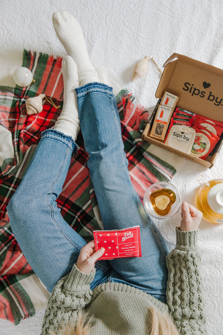 An overhead view of a person in jeans and a sweater holding a cup of tea while sitting on a white bedspread with a green and red tartan blanket and white ornaments scattered around. A box from SIPS BY, full of assorted bags of tea, sits open next to them. 