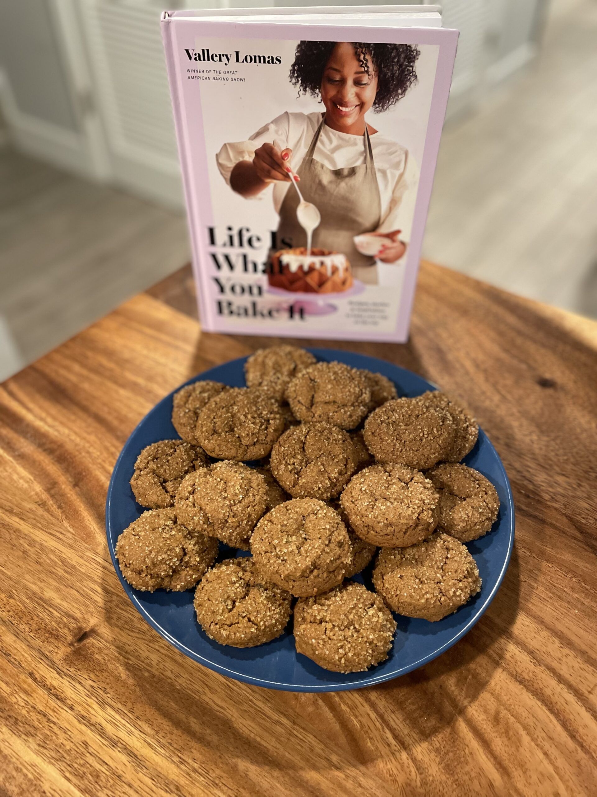 Thick gingerbread cookies covered in coarse sugar on a blue plate beside Vallery Lomas' cookbook Life Is What You Bake It