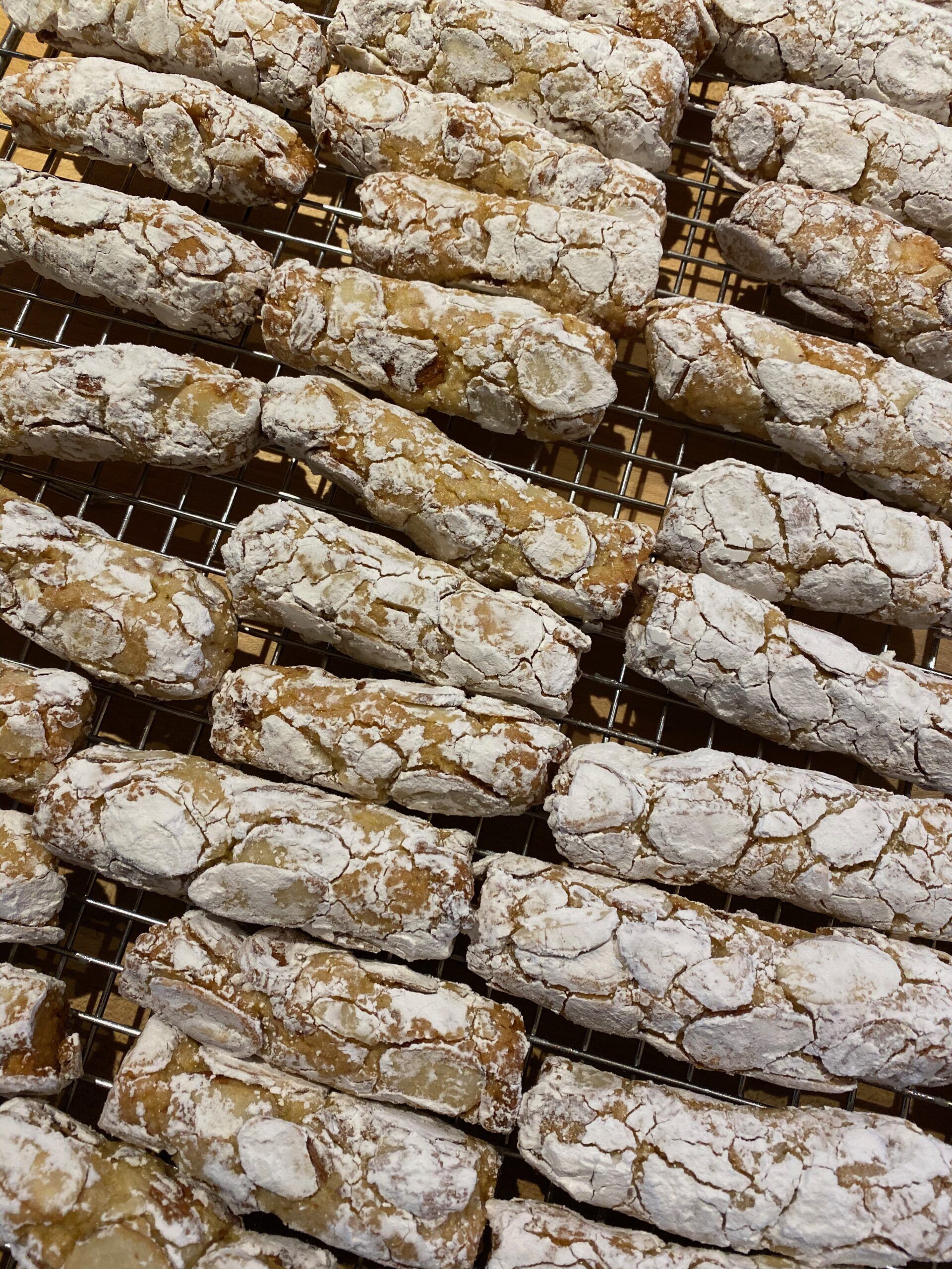 Small, oblong, cylinder shaped cookies, covered in flaked almonds and powdered sugar, sit on a metal cooling rack.