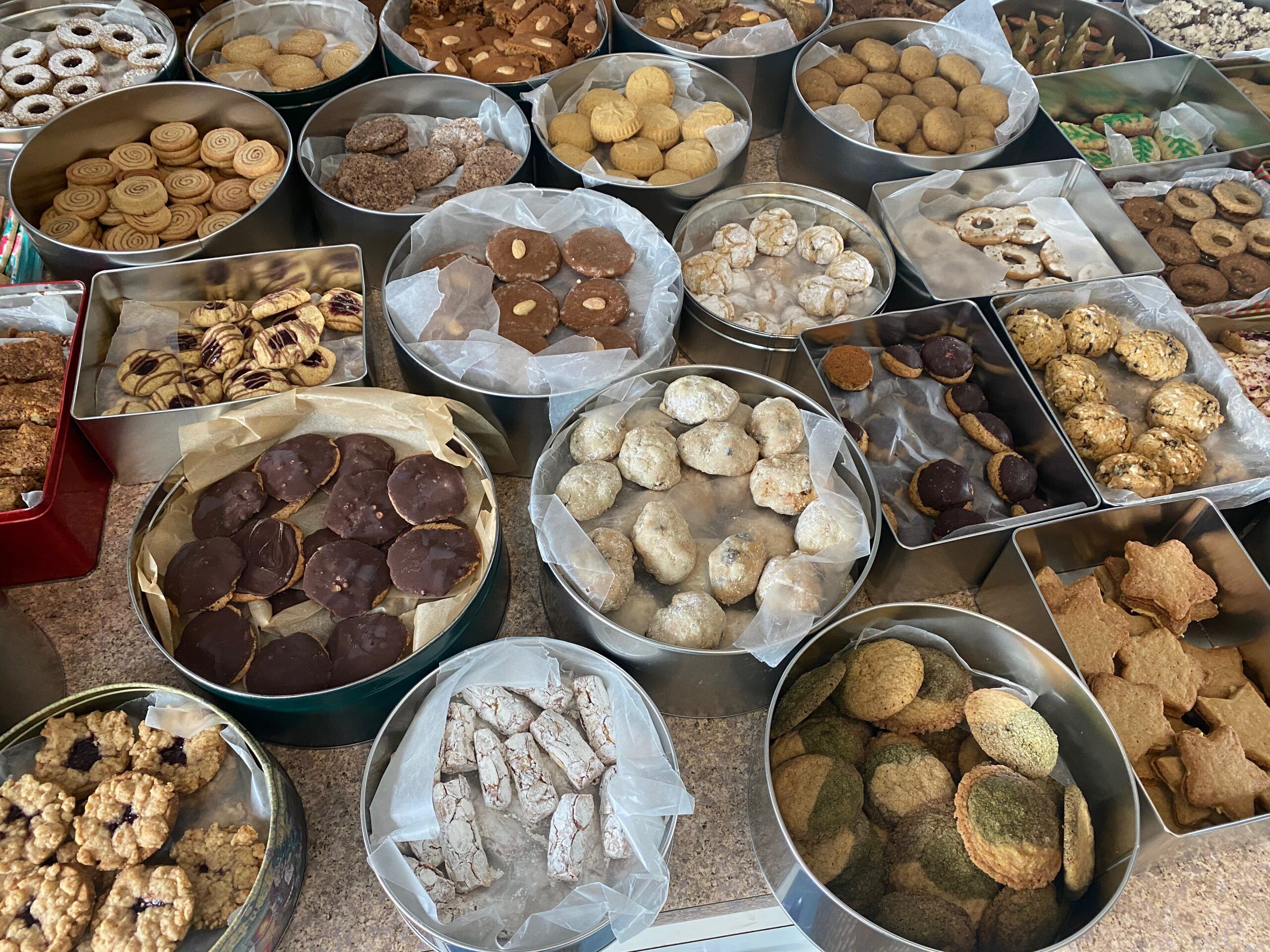 An array of cookies in metal tins on a counter. They cookies are all different shapes and sizes, including jam filled circles, star-shaped cookies, pinwheel cookies, and chocolate cookies. Photo taken by me.