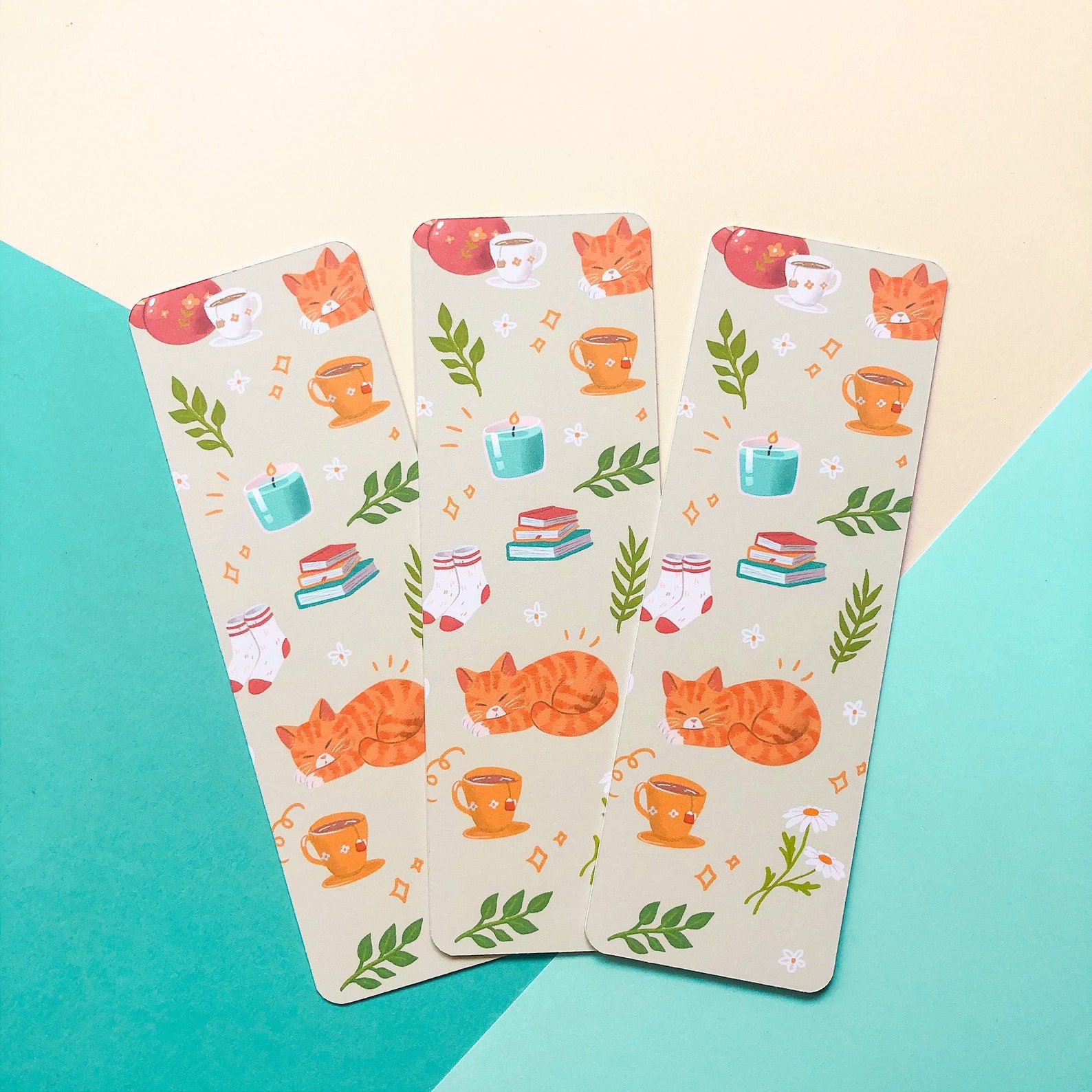 Image of three bookmarks on a teal background. The bookmarks are light  brown and feature a tabby cat, flowers, socks, and tea. 