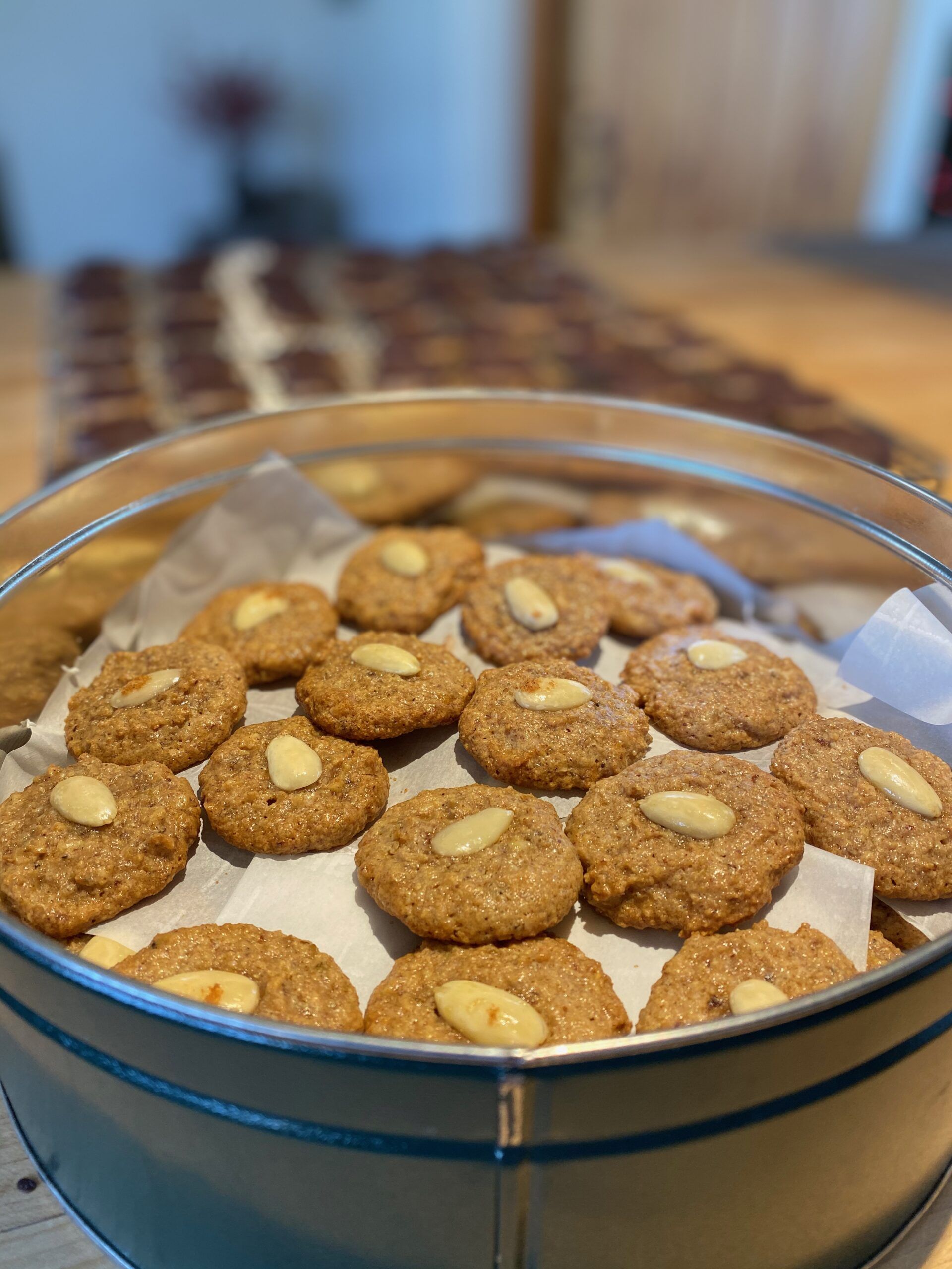 A tin of round golden cookies with an almond in the center of each. A tray of chocolate-covered cookies is blurred out in the background.