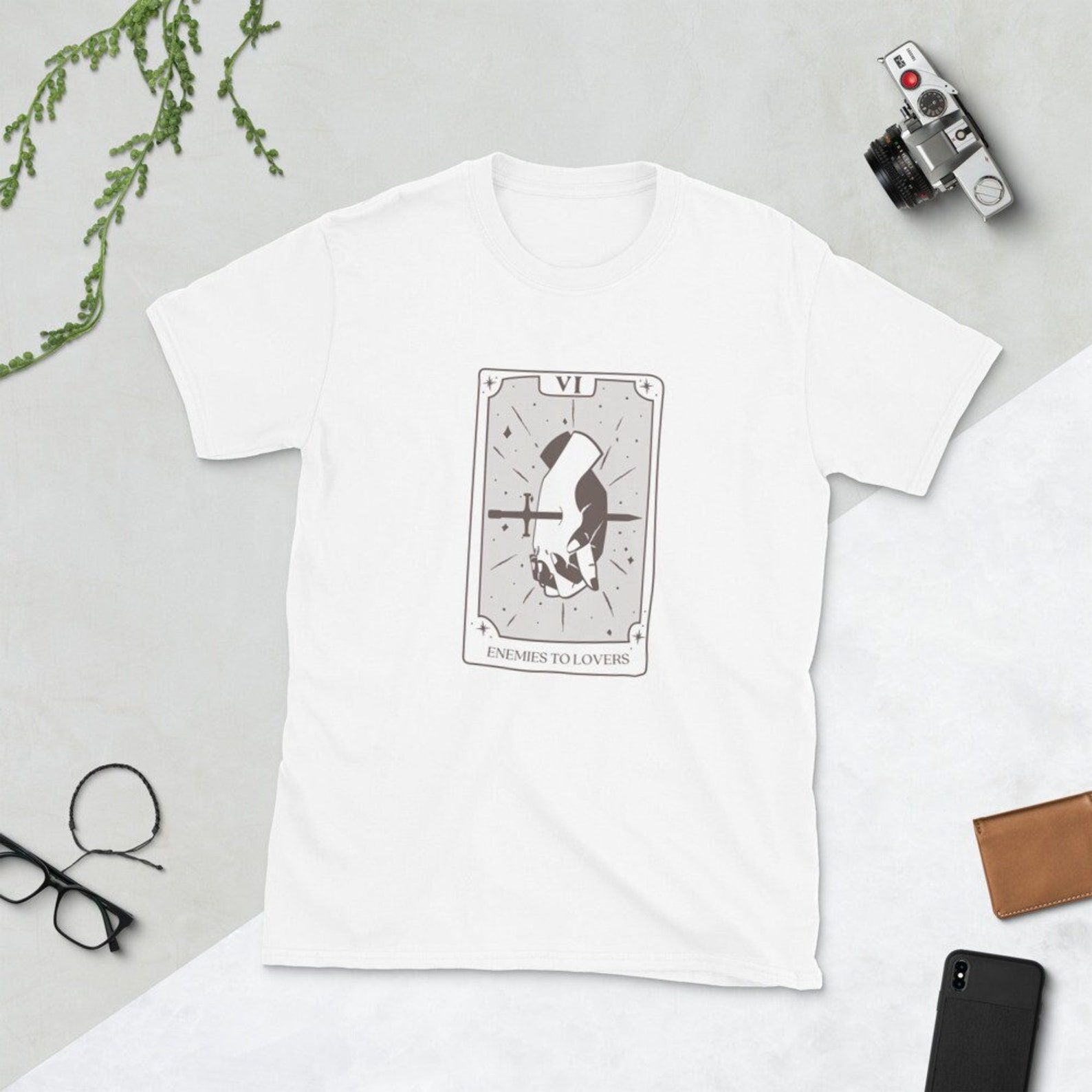 Image of a white t-shirt on a whit background. It has a tarot card in the center with two hands entwined and a sword through them. It's called the enemies to lovers card. 