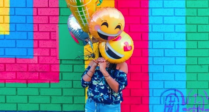 a young woman covers her face with smiley face balloons in front of a colorful wall