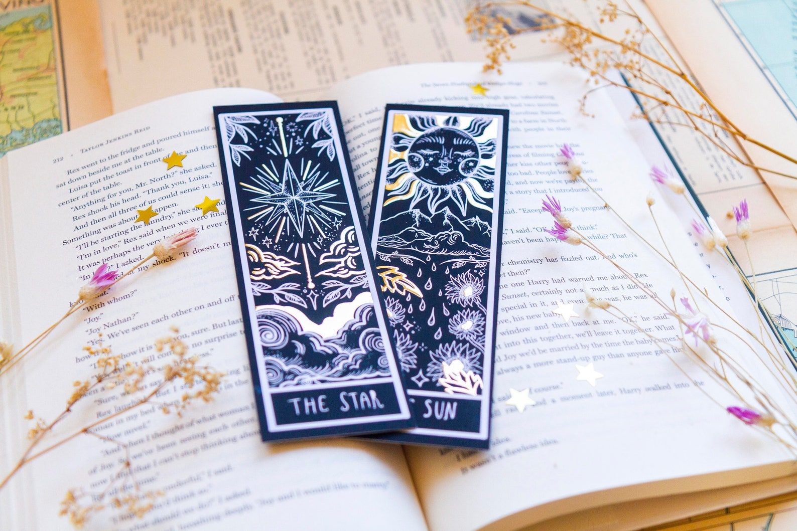 Image of two black bookmarks on top of an open book. The bookmarks have gold foil and the two pictured are a take on The Star card and The Sun card. 