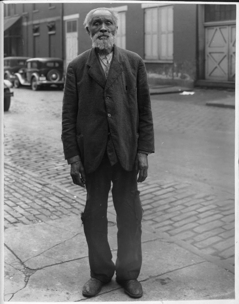 A black and white photograph of Richard Toler, an elderly Black man, standing on a city sidewalk