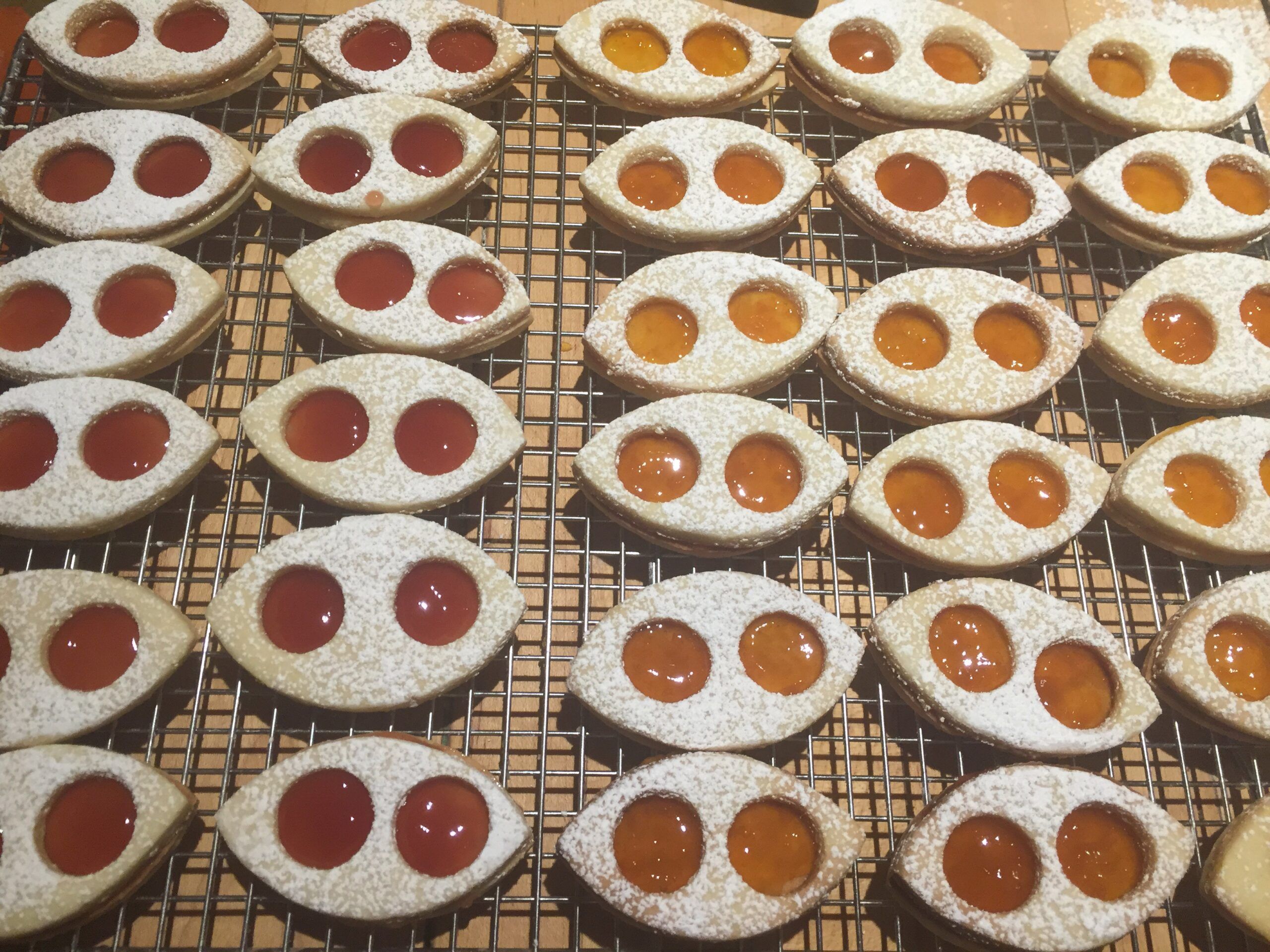 A tray of tear-dropped shaped sandwich cookies, each with two small circles cut out of the tops. Some are filled with red raspberry jam, others with golden apricot jam. Photo taken by me.