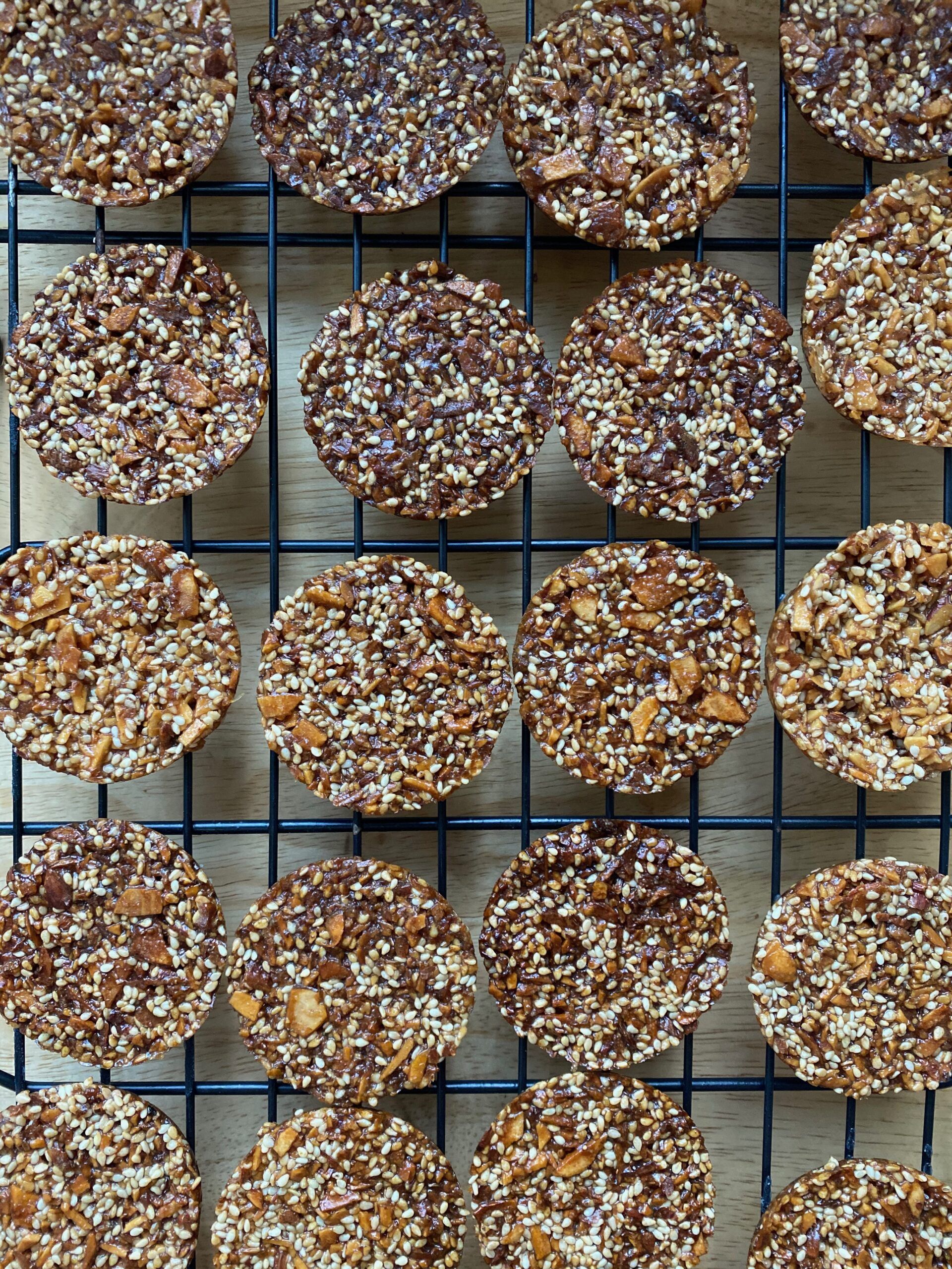 A cooling rack full of round cookies, studded with sesame seeds and chopped almonds. Photo taken by me.