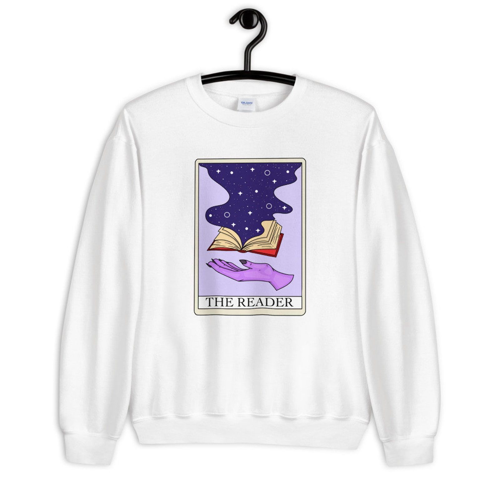 Image of a white sweatshirt with a purple tarot card in the middle. The card features a hand and a book and is titled "The Reader."