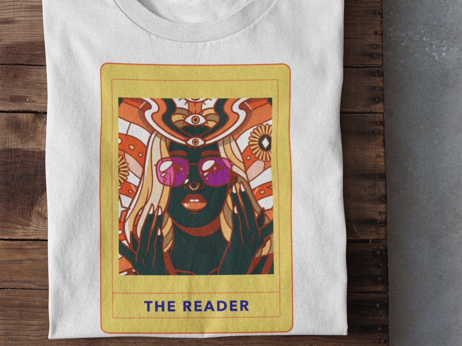 Image of a white tee, featuring "The Reader" card in the middle. The card features a Black skinned person with vintage 70s styling. 