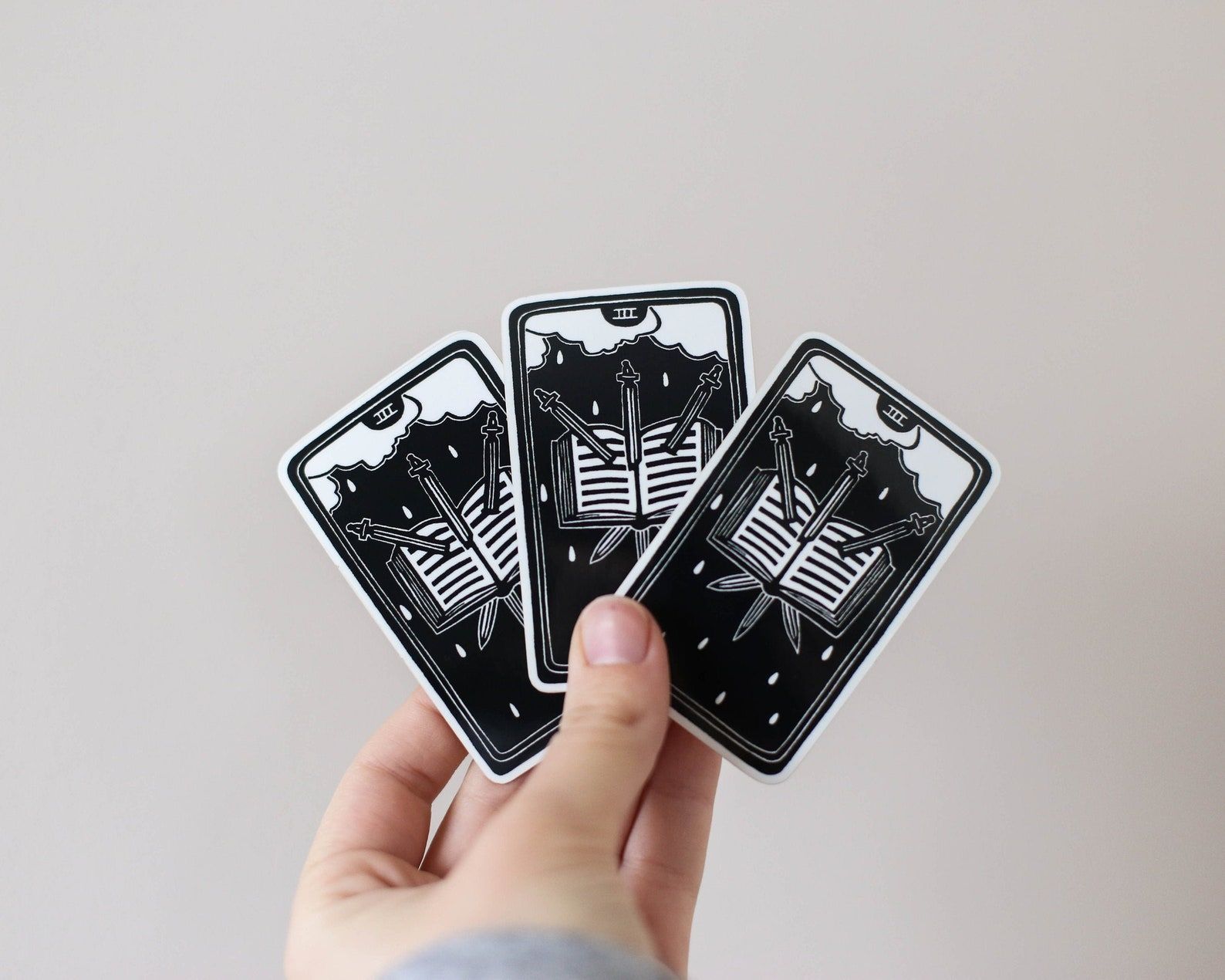 Image of a white hand holding three stickers. Each is in the design of The Three of Swords card, featuring a trio of swords through an open book. The stickers are black with white ink. 