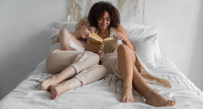 a photo of two women cuddling and reading together in bed. One is Black and one is white