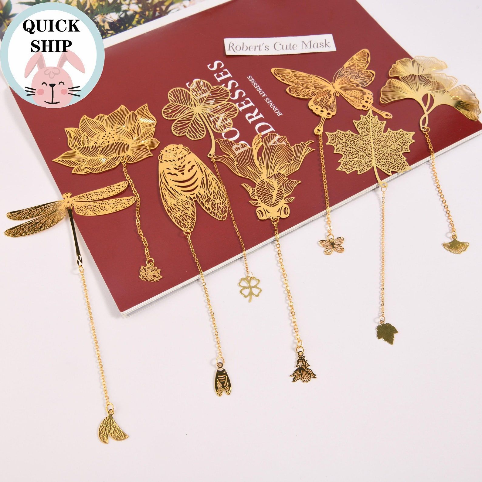 Image of an array of gold metal bookmarks, including a cicada, a lotus, and a gingko leaf. All of them are on a red book cover. 