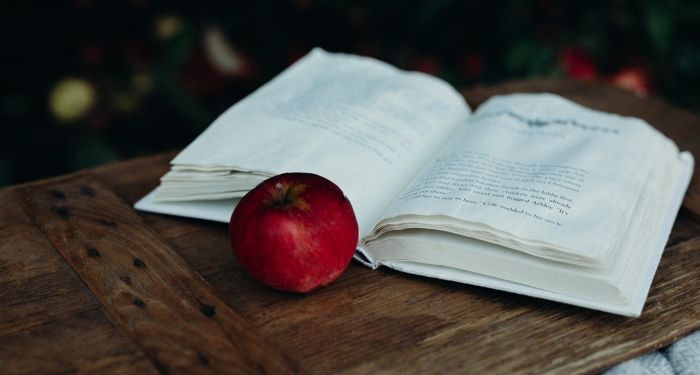 an open book on a wood surface with a red apple next to it