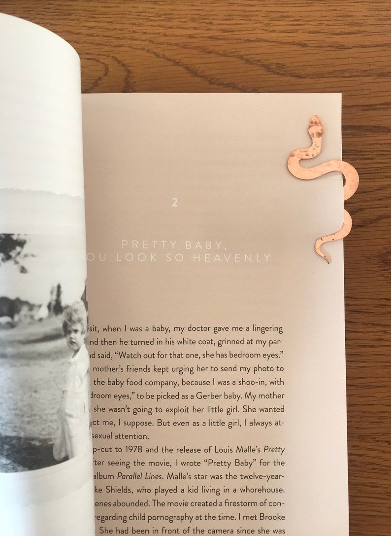 Image of a gold and a rose gold metal bookmark in the shape of a serpent. They're on and beside an open copy of a book.  
