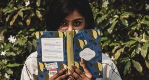 Indian woman holds a book to her face, obscuring her nose and mouth, and also showing her silver fingernails. She's standing in front of a a flowery bush