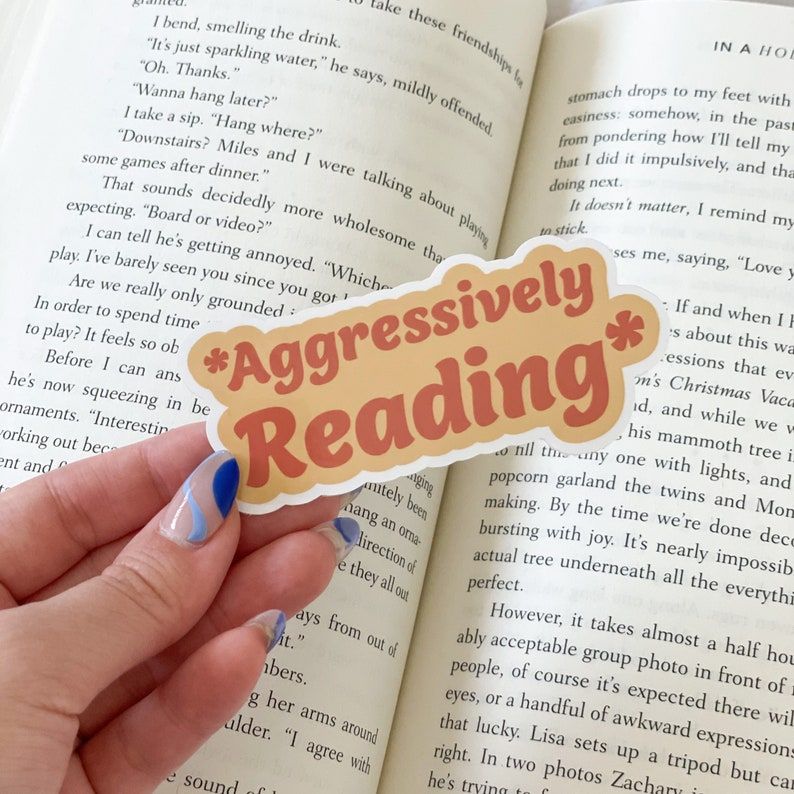 Image of a white hand holding a sticker in front of an open book. The sticker is light brown with pink font. It says *aggressively reading*.