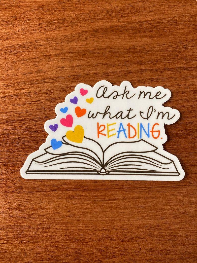 Image of a white sticker on a wood background. The sticker is an open book with the words "ask me what i'm" in black and "reading" in a rainbow of colors. 