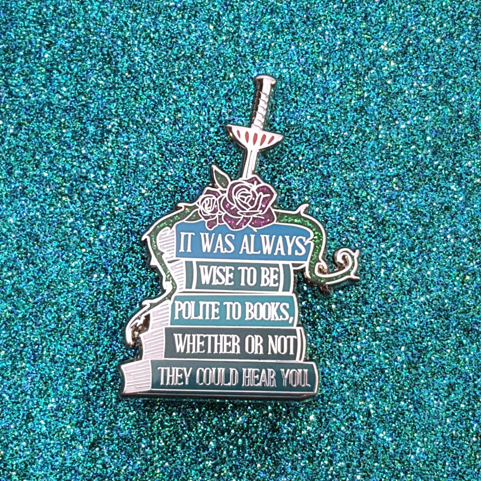 An enamel pin in the shape of a stack of books with the text on spines reading "It's always wise to be polite to books, whether or not they could hear you"