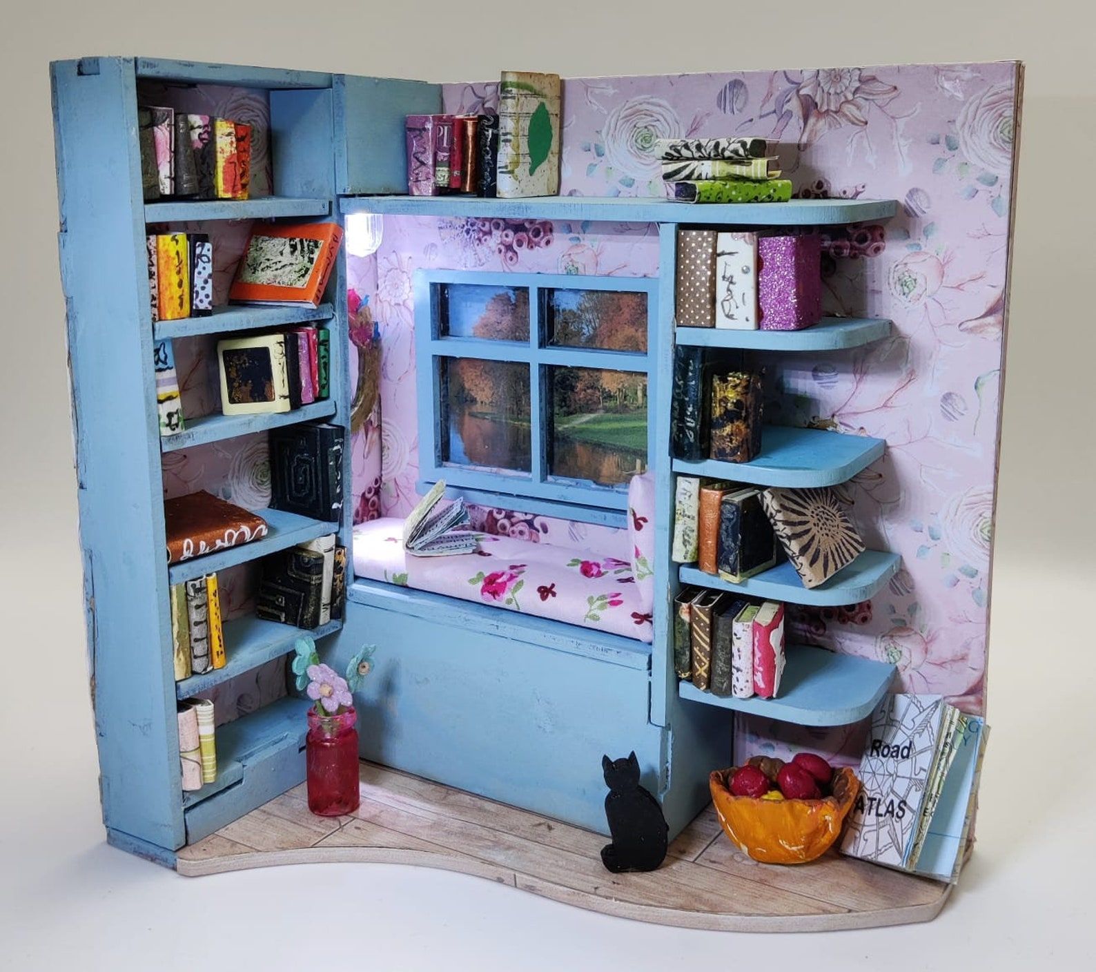 Image of a miniature book nook. it's blue with a window seat, a black cat, and books. 