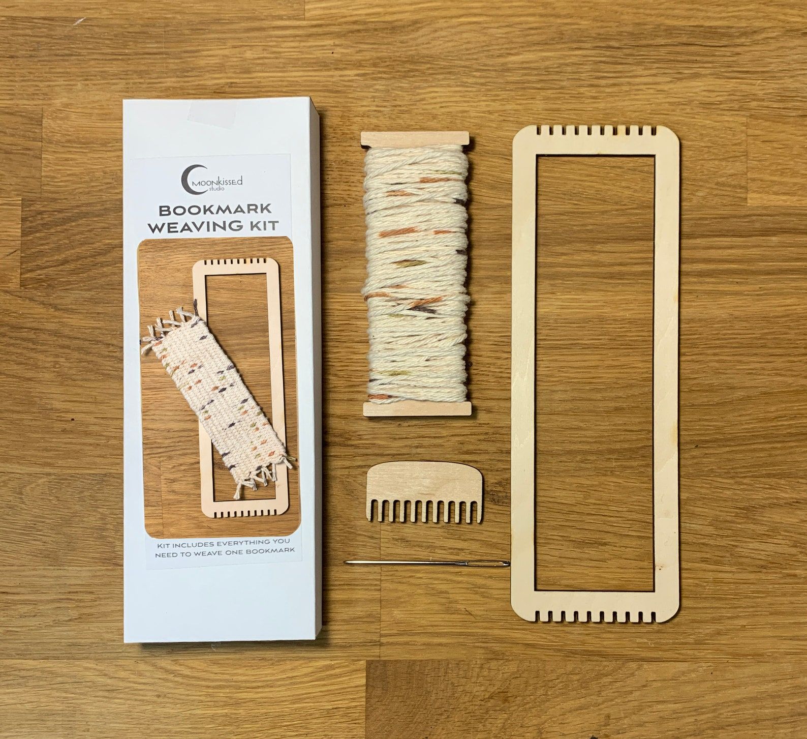 Image of a woven bookmark loom kit, featuring a small comb, loom, and needle. 