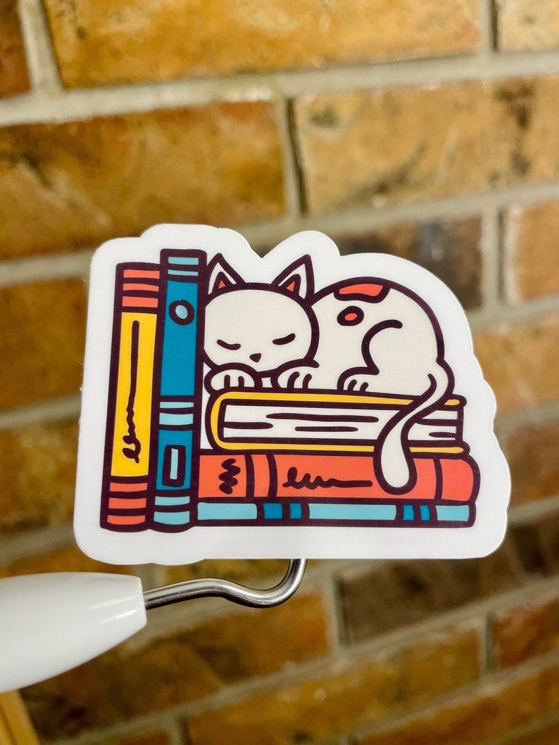 Image of a white cat on a stack of colorful books. The sticker is in front of a brick wall. 