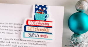 A magnetic bookmark depicting a colorful stack of books and a steaming cup of hot cocoa that reads "Cozy books, hot chocolate, snowy days"