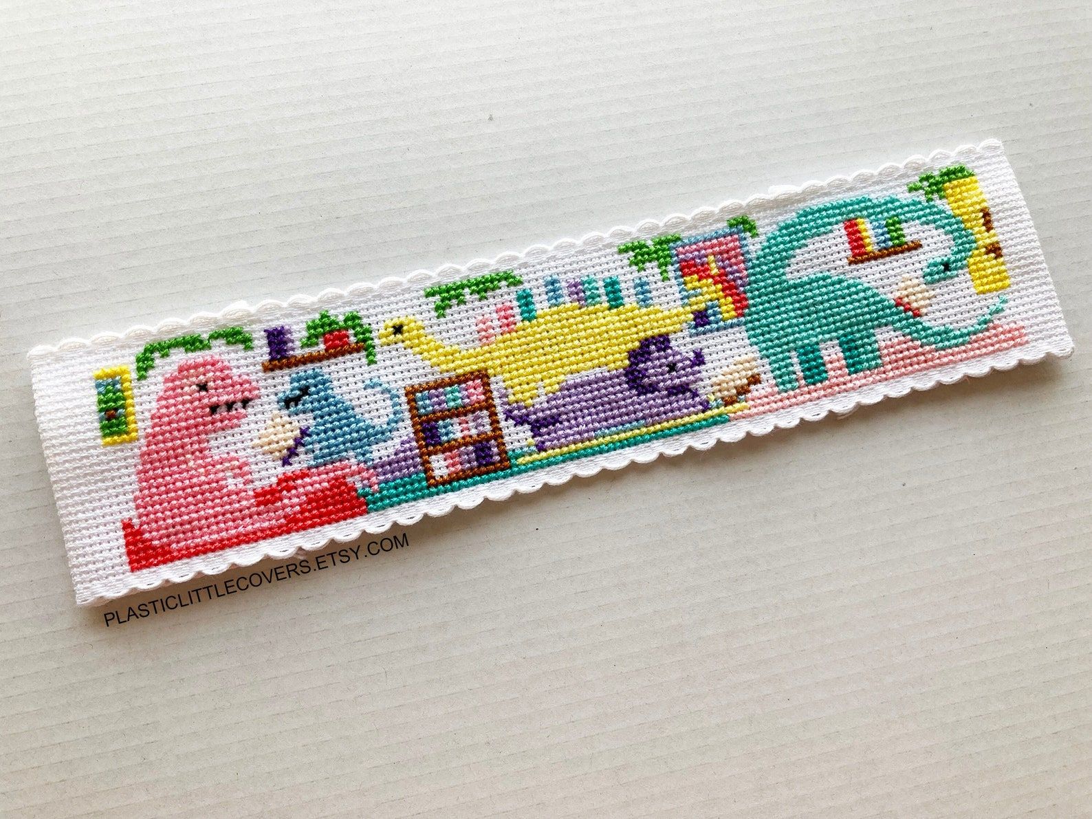 Image of a cross stitch bookmark. it features colorful dinosaurs inside of a library. 
