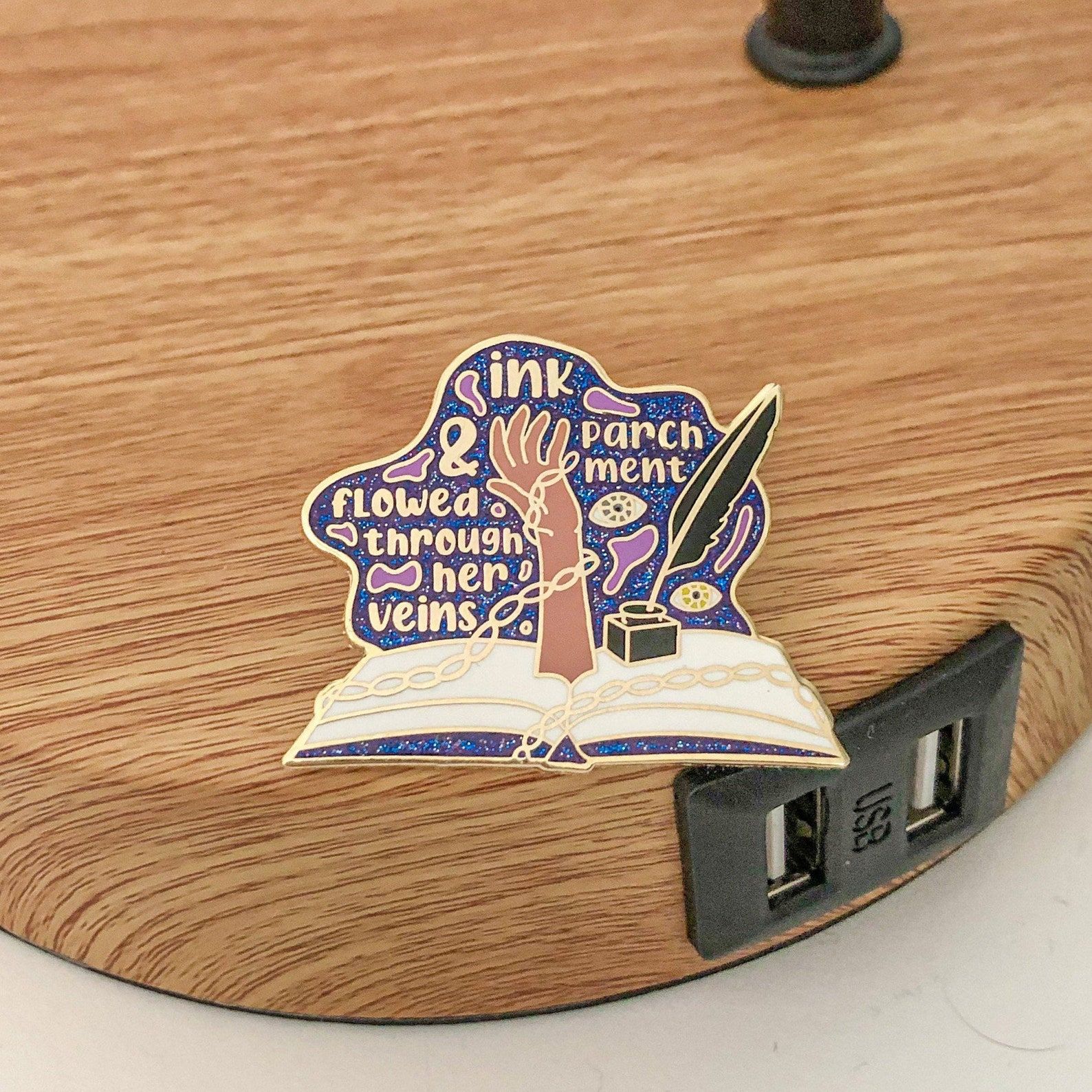 An enamel pin with a hand, quill pen and ink, and the words "ink and parchment flowed through her veins"