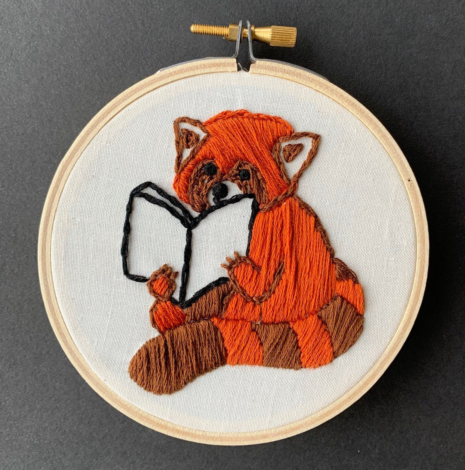 Image of a finished embroidery project, featuring a red panda reading a book. 