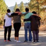 four teens with their backs towards the camera standing on a road in a wooded area with arms linked