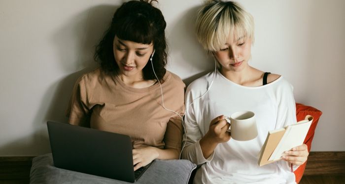 two women sitting on a bed together sharing headphones, with one reading a book and the other on her laptop