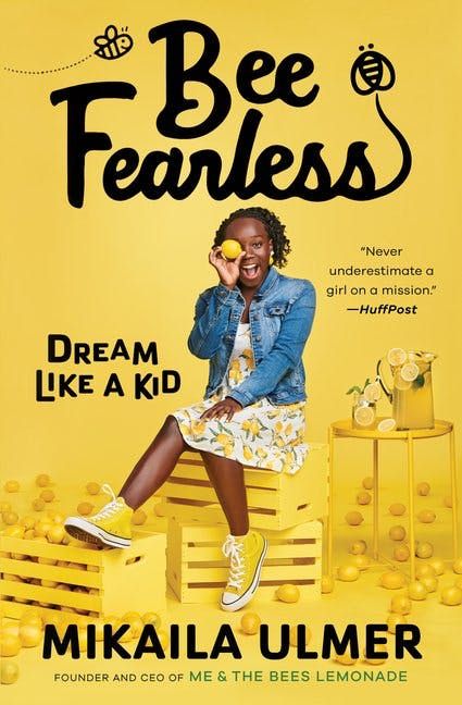 A 15-year-old Black girl in a jean jacket and yellow skirt sits on yellow crates on a yellow background. She is holding a lemon. It is the cover of her book, Bee Fearless - Dream Like A Kid by Mikalia Ulmer.