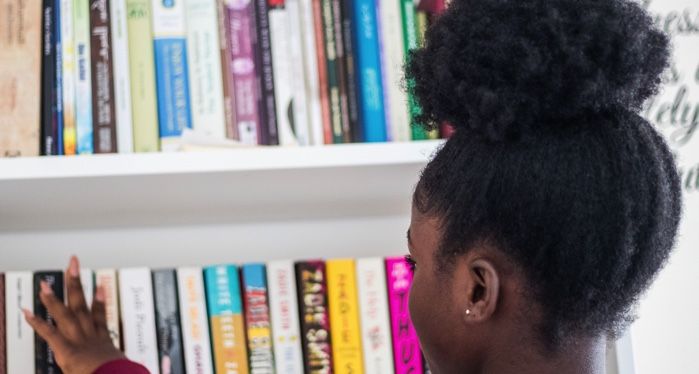 a dark brown-skinned young girl with a big afro puff and burgundy shirt is looking at a bookshelf