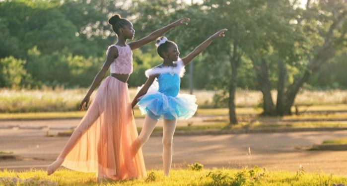 two Black child ballet dancers dressed in pink and blue, one leg outstretched and one arm reaching