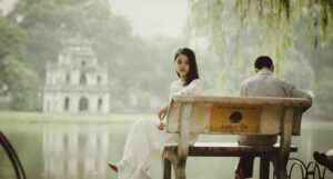 Vietnamese man and woman sitting on bench. The woman is turned away from the man and seems to be deep in thought. The bench is by a pond with a historic building in the distance