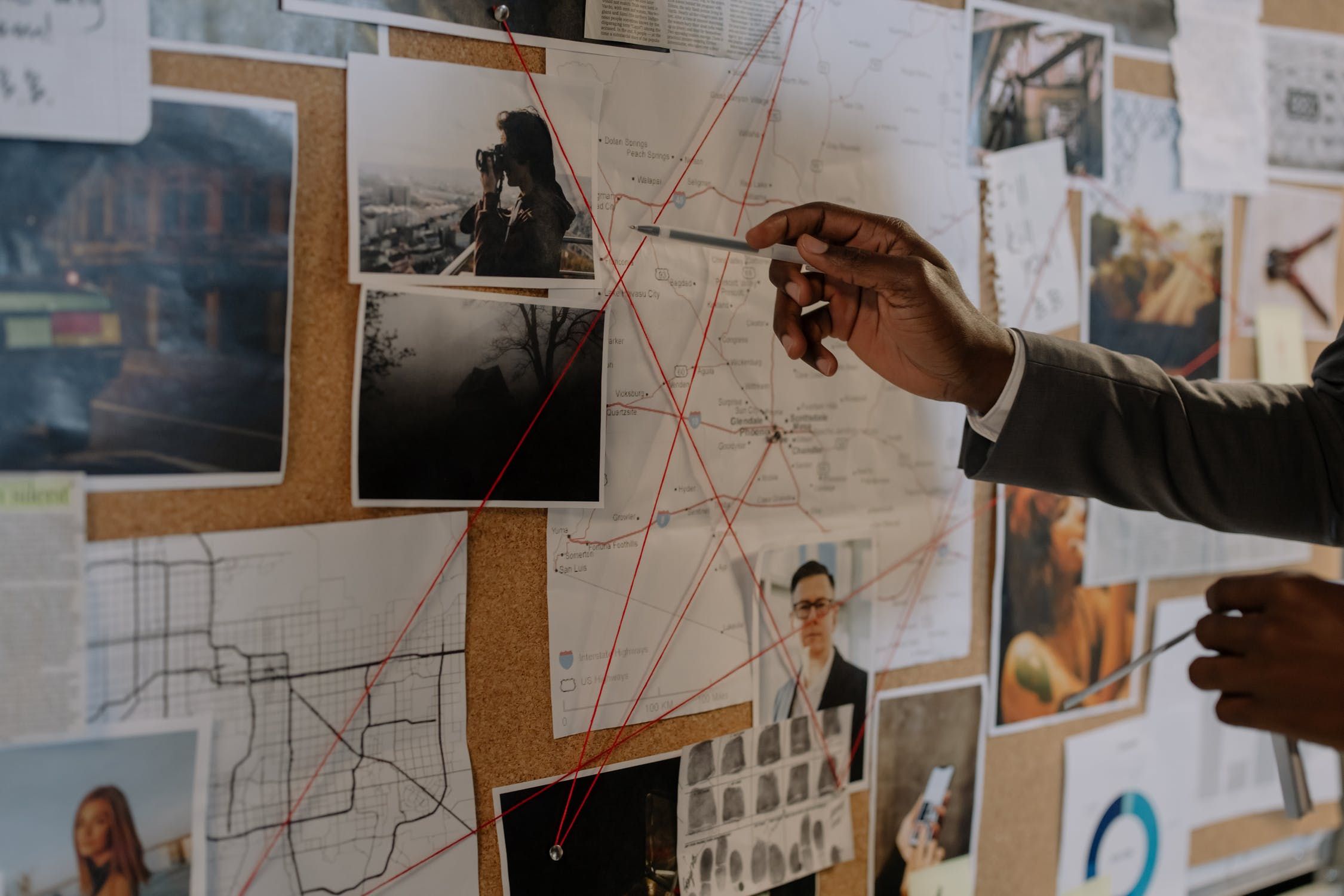 Image shows a pinboard covered in photos, maps and documents, with red strings connecting them. A man's hands and right arm are visible; he is pointing at the central photo with a pen. 