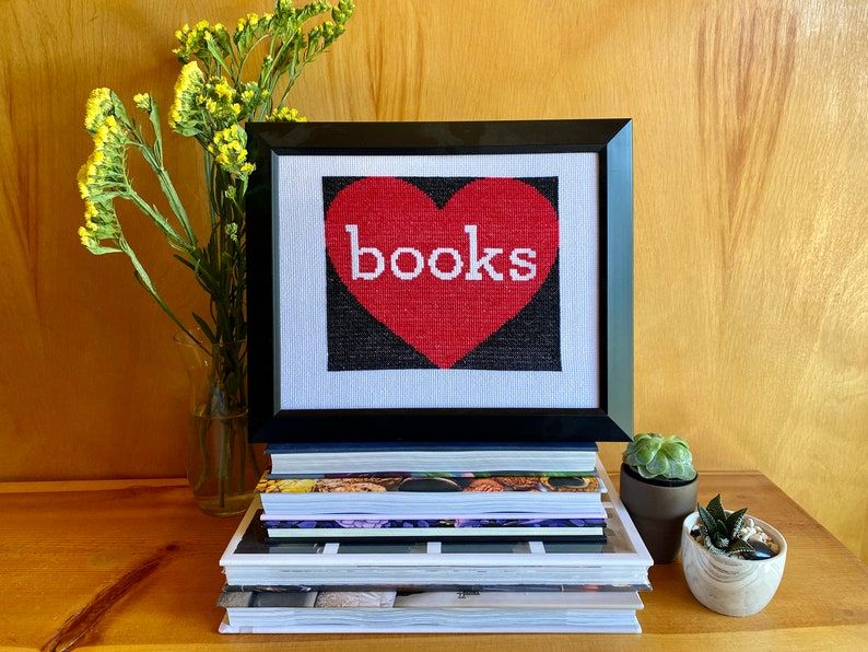 Image of a finished cross stitch on top of some books. The cross stitch is a big heart with the word "books" inside. 