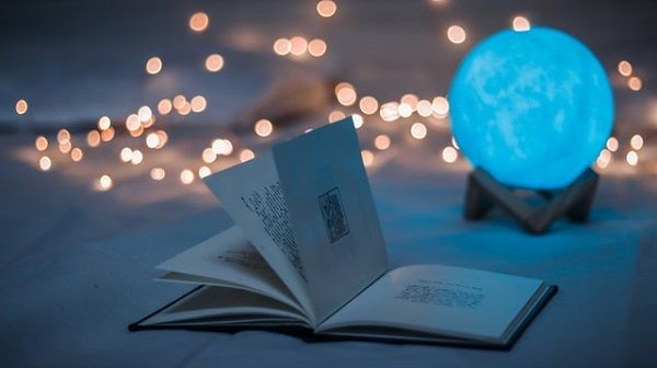 An open book with a moon light creating a soft blue glow with twinkle lights in the background