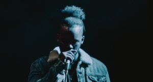 a person with Black skin wearing a denim jacket speaking into a microphone in low light