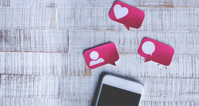 an iPhone on a table. Above it are red paper cut outs that look like social media notifications or icons. There's one in the shape of a person, a heart and a comment box.