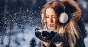 pale skinned woman blowing snow outside in a coat and earmuffs