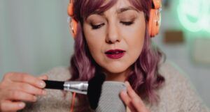 pale-skinned woman with light purple hair is brushing microphone with a makeup brush while wearing orange headphones