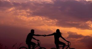 two people riding bicycles; the person with long hair reaches back to grab the other person's hand