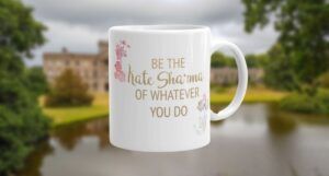 Kate Sharma mug with text: "Be the Kate Sharma of whatever you do." Mug is imposed against a blurred background of a large lakefront estate