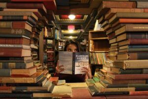 lightly tanned-skin woman sits behind a lot stacked books; she's holding a book so that only her eyes are visible