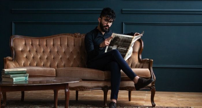light-skinned man with dark hair and full, dark beard is sitting on a settee reading a newspaper with one leg crossed over the other